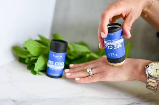 Person holding a container of Trueceuticals Bee Cool on a marble surface with green leaves.