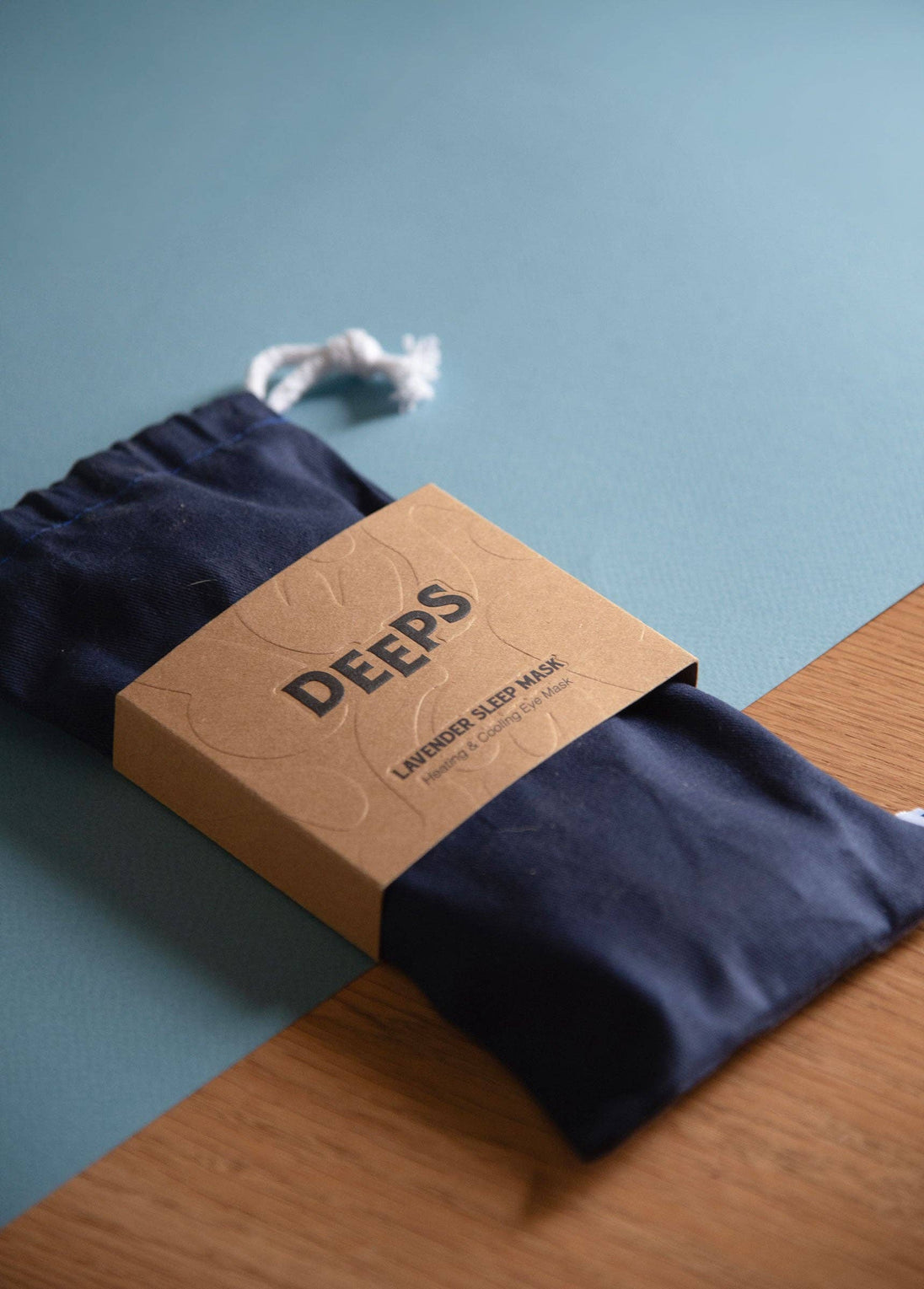 Navy blue sleep mask with 'DEEPS' branded box on a wooden surface and blue background