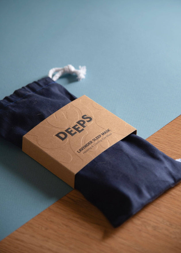 Navy blue sleep mask with 'DEEPS' branded box on a wooden surface and blue background
